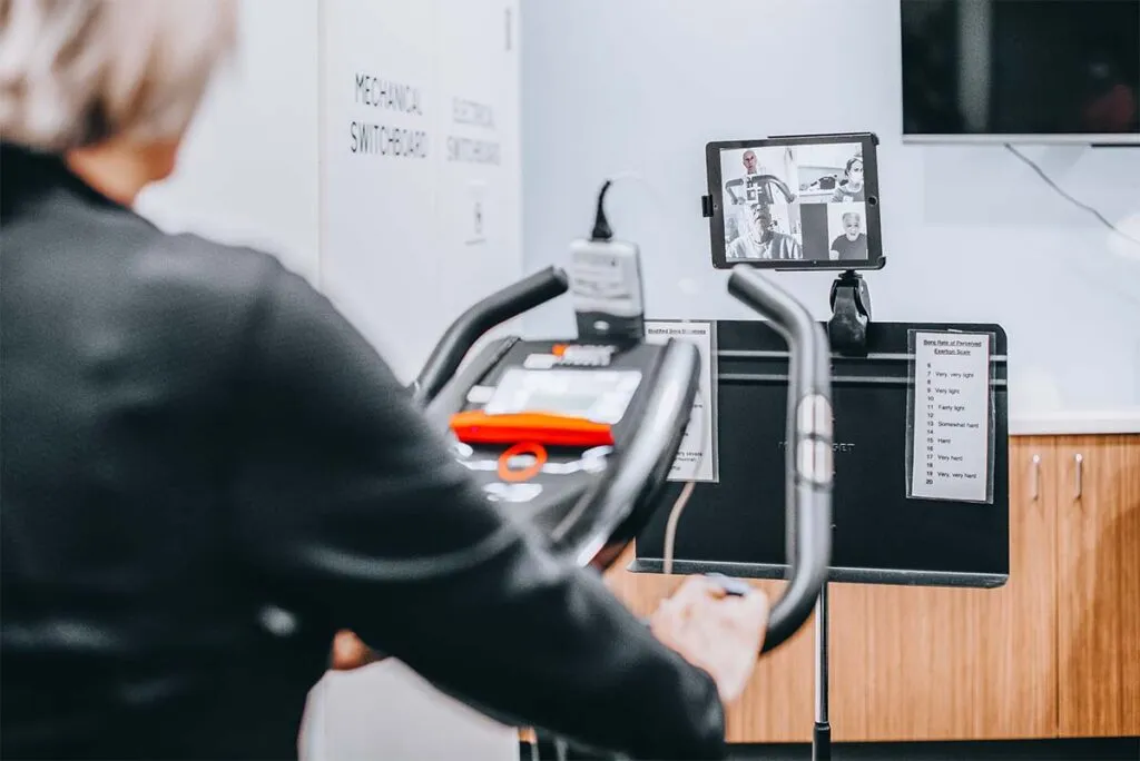A participant exercises on a step-through bike at home, with a tablet on a stand showing a videoconference and a Borg perceived-exertion scale visible alongside.